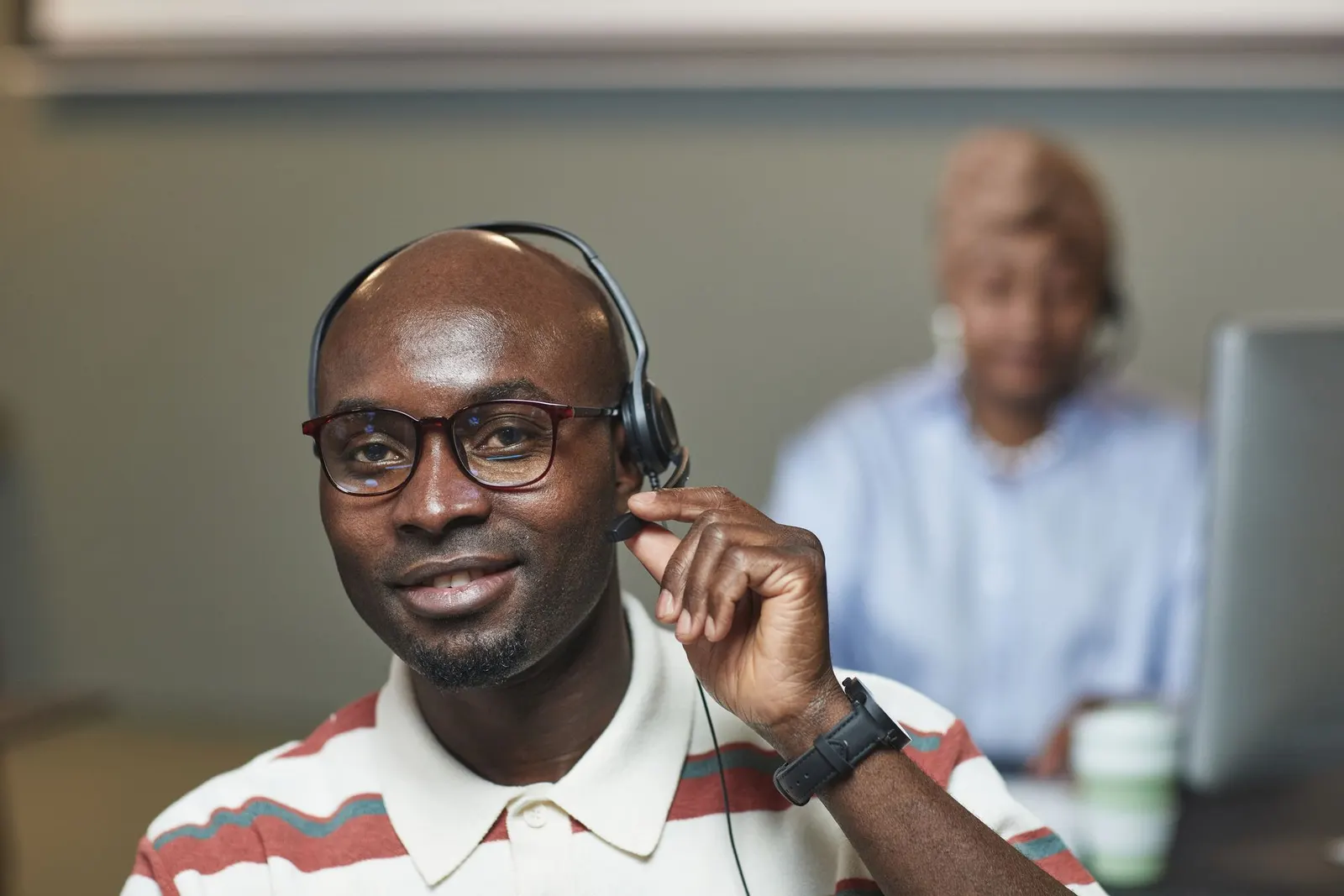 A smiling man wearing a headset and glasses, talking to a customer at his desk.